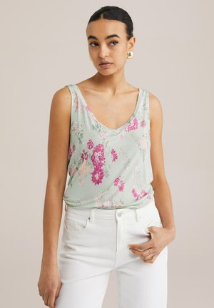 Floral tank top in light green with pink and white flowers; features a V-neck and relaxed fit, paired with white high-waisted jeans.