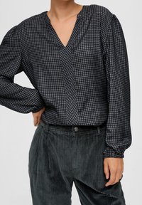 Blouse à motif pied-de-poule noir et gris avec un décolleté en V, des manches longues et des poignets élastiqués, associée à un pantalon en velours côtelé texturé de couleur vert foncé.