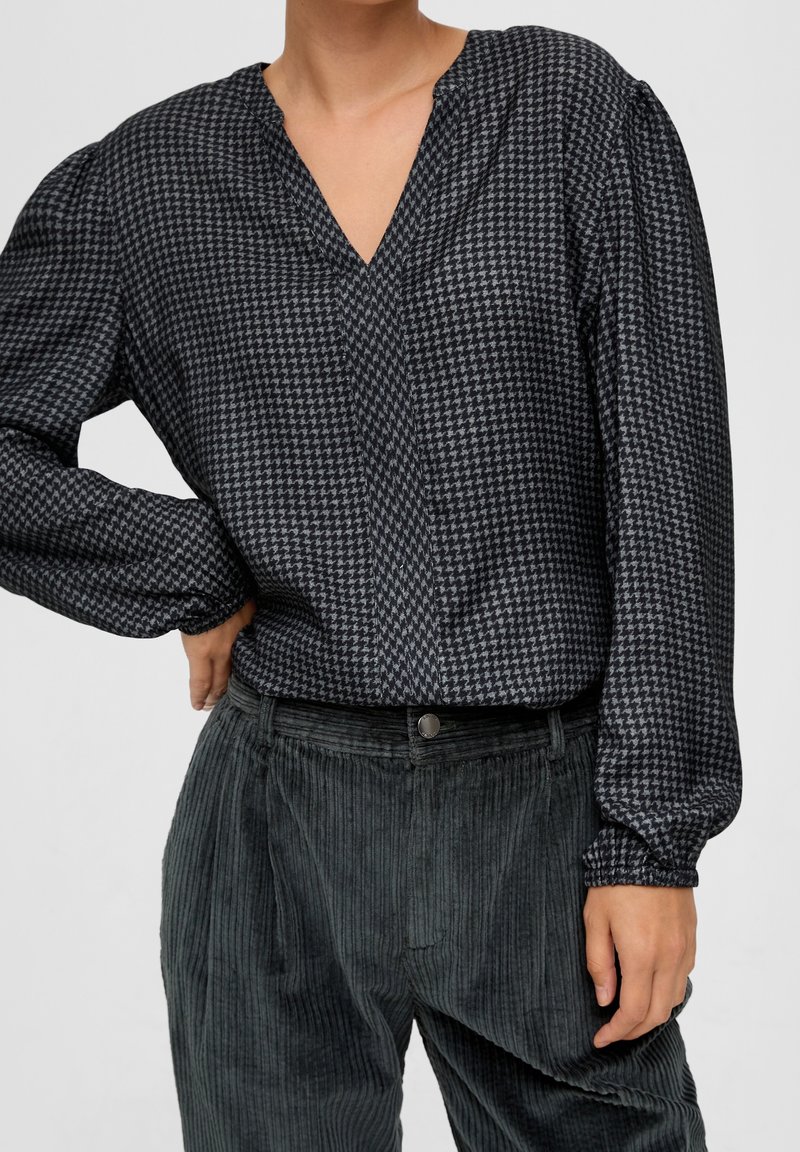 Blouse à motif pied-de-poule noir et gris avec un décolleté en V, des manches longues et des poignets élastiqués, associée à un pantalon en velours côtelé texturé de couleur vert foncé.