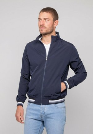Homme portant une veste zippée bleu marine avec des poignets rayés blancs et gris et un jean bleu clair, regardant vers sa gauche avec la main dans la poche.