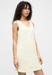 Γυναίκα με κατσαρά μαλλιά, φορόντας μία αμάνικη κρεμ φόρεμα μέχρι το γόνατο με υφασμάτινες λουλουδένιες εφαρμογές, στέκεται μπροστά σε απλό φόντο.