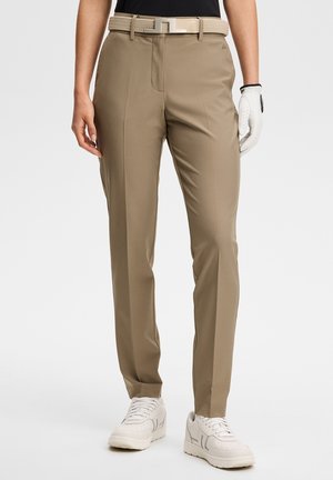 Persoon die beige op maat gemaakte broek met plooi draagt, beige geweven riem, witte golfhandschoen aan de rechterhand en witte sneakers.