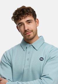 Lyseblå langærmet polo med krave, tre knapper og en marineblå logo-patch på venstre side. Blød tekstur.