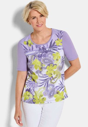 Blonde Frau trägt ein lavendelfarbenes Kurzarmoberteil mit gelben und violetten Blumen- und Paisleymustern, kombiniert mit weißen Hosen.