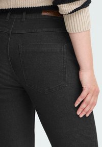 Femme portant un jean en denim noir avec une poche arrière visible et une manche de pull en tricot rayé beige et noir.