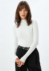 Valkoinen ribbattua turtleneck-pitkähihainen paita, jossa on istuva muotoilu, nilkkakaulus ja peukaloaukot, yhdistettynä korkeavyötäröisiin mustiin housuihin.