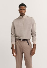 Beige Strickpullover mit Stehkragen und halbem Reißverschluss, kombiniert mit hellbraunen Hosen und einem geflochtenen Gürtel. Strukturiertes Muster sichtbar.