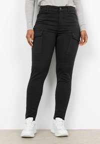 Černé vysoké cargo kalhoty s bočními kapsami, slim fit a hladkým povrchem, doplněné bílými sportovními botami.
