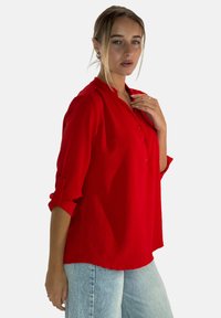 Blusa roja texturizada con botones en la parte delantera y mangas remangadas, combinada con jeans azul claro. Cuello destacado y diseño de ajuste relajado.
