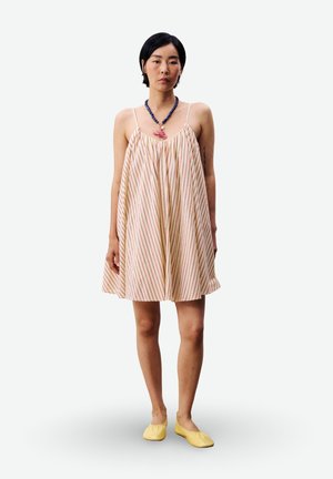 Femme portant une robe ample et sans manches de couleur crème avec de fines rayures rouges, des ballerines jaunes, et un collier épais bleu et rose, debout devant un fond blanc.