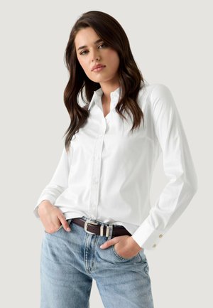 Femme aux longs cheveux bruns portant une chemise blanche boutonnée rentrée dans un jean bleu clair, les mains dans les poches, debout devant un fond uni.