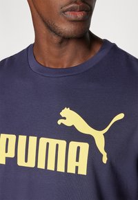 T-shirt en coton bleu marine avec un grand logo "PUMA" jaune et un graphique de puma bondissant au-dessus. Encolure ras du cou et manches courtes.