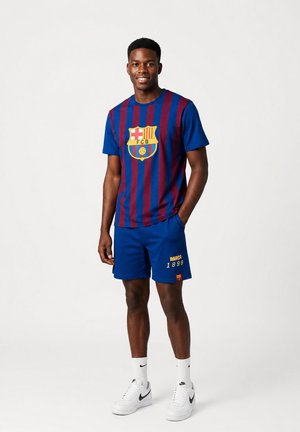 Młody mężczyzna w niebiesko-czerwonej pasiastą koszulką FC Barcelona i niebieskich szortach z białymi butami Nike, stojący na jednolitym tle.