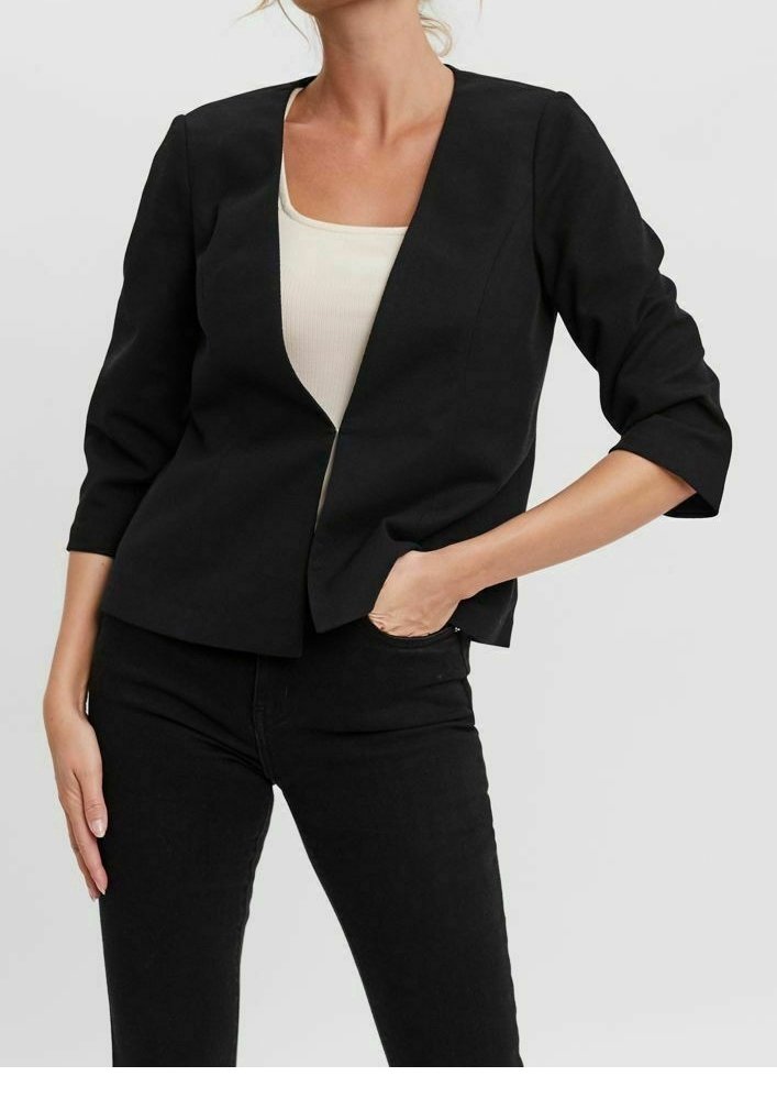 Femme portant un blazer noir à manches trois-quarts sur un haut blanc, associé à un pantalon noir, une main dans la poche, debout.