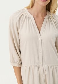 Robe beige clair avec un décolleté en V, boutons sur le devant et manches bouffantes trois-quarts. Comprend des détails froncés au niveau du décolleté et de la taille.