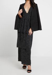 Robe noire à pois avec un décolleté en V profond et un peplum superposé, assortie à un blazer noir ajusté et des sandales noires à lanières.