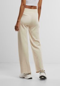 Pantaloni a gamba larga beige realizzati in tessuto morbido e texturizzato; vita alta; indossati con scarpe da ginnastica bianche e un top corto, visti da dietro.