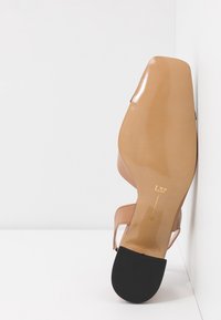 Sandale à talons beige avec une finition laquée brillante, orteil ouvert et fine sangle de cheville. Semelle en caoutchouc noire avec des motifs en relief au fond.