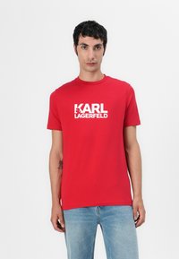 Raudona medvilninė marškinė su trumpomis rankovėmis ir apvaliu kaklu, su ryškiu baltu "KARL LAGERFELD" logotipo atvaizdu priekyje.