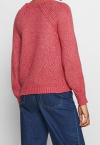 Pletený svetr v růžové barvě s texturovaným vzorem, spadlými rameny a žebrovanými manžetami a lemem, ve spojení s modrými džínami.