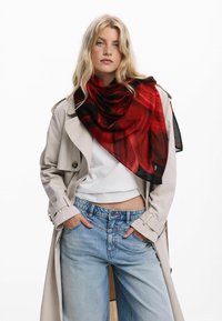 Sciarpa leggera con motivo rosso e nero, drappeggiata elegantemente attorno al collo, in contrasto con un trench beige e jeans.