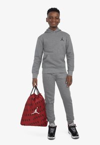 Sivi pamučni dukserica i trenirke s crnim Jumpman logom, u kombinaciji s crno-crvenim tenisicama; drži crvenu torbu s utezima na crnom slovu.