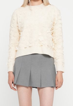 Femme portant un pull en maille crème texturée avec des appliqués floraux et une mini-jupe plissée grise taille haute.