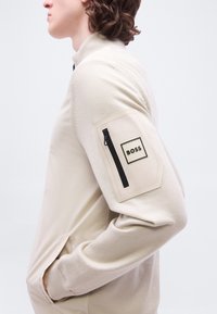 Προφίλ ατόμου που φοράει μπεζ πουλόβερ με τσεπάκι με φερμουάρ και λογότυπο BOSS στην πάνω πλευρά του μπράτσου, με τα χέρια στις πλαϊνές τσέπες.