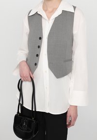 Persona vistiendo una camisa blanca abotonada y un chaleco gris, sosteniendo un bolso de cuero negro con correas largas sobre un fondo liso.