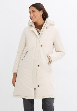 Giacca puffer beige con zip frontale, due tasche frontali e cappuccio. Texture morbida e trapuntata con bottoni a contrasto e dettagli di cucitura.