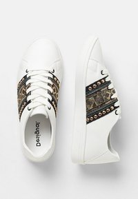 Zapatillas blancas de piso bajo con rayas laterales de patrón negro y dorado y tachuelas doradas, vistas desde ángulos superior y lateral.