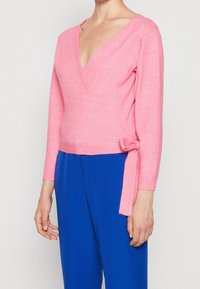 Pink wrap sweater with a deep V-neck, long sleeves, and a side tie. Soft texture and fitted design. Paired with blue trousers.