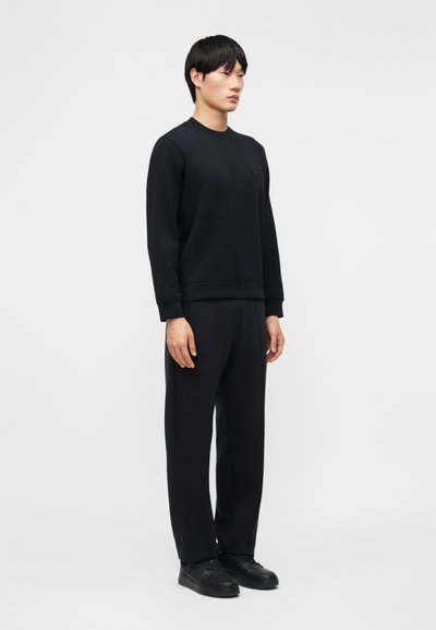 Άνδρας στέκεται φορώντας μια ολόμαυρη στολή με μακρυμάνικο φούτερ, παντελόνι με ίσια γραμμή και μαύρα αθλητικά παπούτσια σε λευκό φόντο.