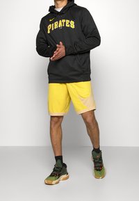 Felpa nera con la scritta "PIRATES" in giallo, abbinata a pantaloni corti gialli con un accento beige. Sneakers verdi con dettagli rossi e calze nere.