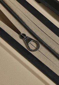 Chiusura con zip in tessuto beige con tirazip metallico, circondata da accenti neri e un motivo testurizzato sul materiale, che offre un design elegante.
