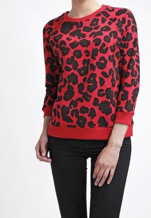 Femme portant un sweat-shirt rouge à manches longues avec des taches léopard noires et un pantalon noir, debout devant un fond blanc uni.