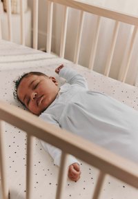 Wit slaapkleedje op een zuigeling die ligt op een beige matras met vlekjes in een houten wieg met een natuurlijke afwerking.