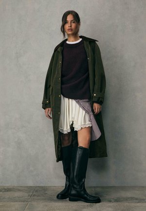 Olive zielony długi płaszcz na ciemnym swetrze, biała spódnica z koronkowym wykończeniem i czarne wysokie kozaki. Teksturowane tkaniny i warstwowy design.