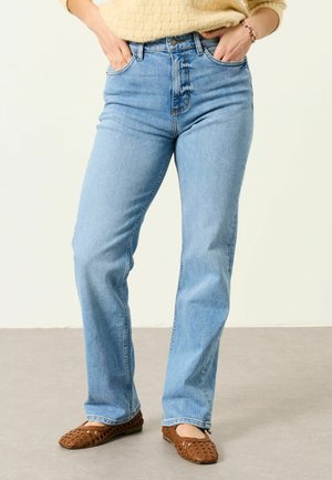 Person wearing light blue straight-leg jeans, beige textured sweater, and brown woven flats, standing with hands in pockets.