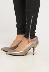 Zapatos de tacón alto metálicos dorados con punta afilada y un acabado texturizado. Combinados con pantalones negros ajustados que presentan un detalle de cremallera plateada.