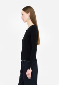Mujer con cabello largo y liso, lleva un cárdigan negro con ribete plateado y pantalones negros a juego, de pie de perfil contra un fondo blanco.