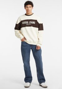 Sudadera crema y marrón con el texto "GUESS JEANS", combinada con unos vaqueros de denim azul holgados y zapatillas blancas. Tejido suave, diseño casual.