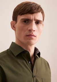 Camicia verde oliva con colletto a contrasto e motivo. Realizzata in tessuto liscio, presenta una tasca sul petto e una patta con bottoni.