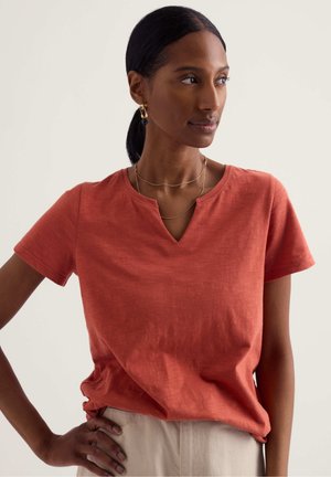 Vrouw met donker haar in een staart, draagt een roestkleurige top met korte mouwen, beige broek, gouden oorbellen en gelaagde gouden kettingen, kijkt naar rechts.