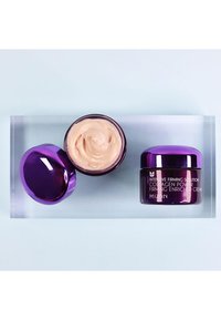 Lila Cremedose mit glänzendem Deckel, die eine hellbeige Creme enthält. Neben ihr steht eine ähnliche, kleinere Dose mit Etikettendesign. Auf einer klaren Oberfläche platziert.