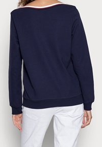 Námořnická modrá mikina s širokým výstřihem a kontrastními bílými a červenými pruhy na zádech; žebrované manžety a lem. Měkká, hladká textura.