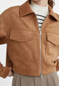 Femme portant une veste en suède marron raccourcie avec une fermeture éclair à l'avant, des poches à rabat, sur un pull blanc côtelé à rayures noires.