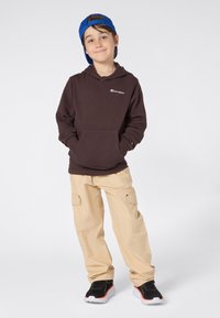 Brun hette med frontlomme, logo på brystet, kombinert med beige cargo-bukser. Har på seg en blå caps og svarte treningstøvler med hvite såler.
