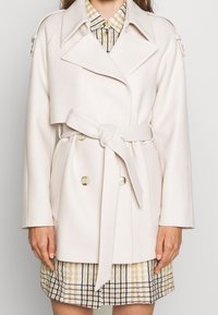 Jasnobeżowy płaszcz typu wrap z szerokimi klapami, podwójnymi guzikami i wiązanym w pasie. Teksturowana tkanina, o długości do bioder, w zestawieniu z sukienką w kratę.