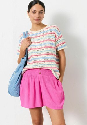 Žena oblečená v prúžkovanom krátkom tričku a jasne ružovej sukni, držiaca modrú tašku na rameno, stojaca pred bielou stenou.